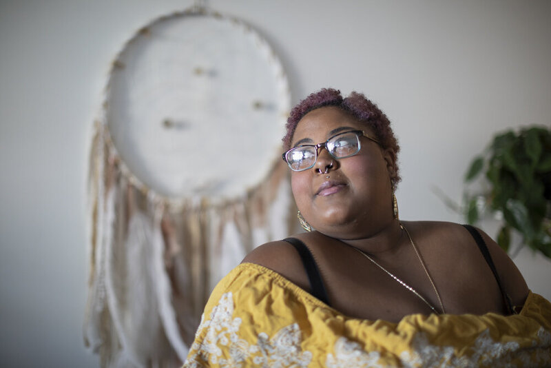 This image frames a black woman, looking over her shoulder past the viewer. She has short, curly pink hair and a septum piercing, and she is wearing a textured yellow and white top with gold jewelry. A hanging woven art piece is visible in the background of the image.