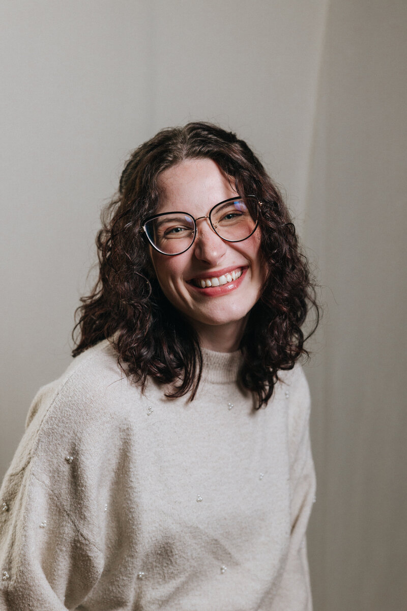 Smiling woman with curly hair and glasses during a professional studio headshot session, wearing a cream sweater.