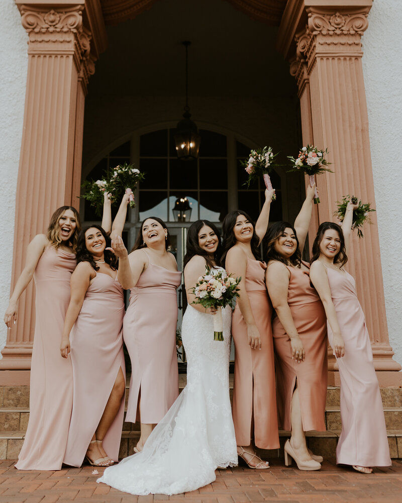 Bride standing in the middle of six bridesmaids who are wearing peach and pink dresses and holding bouquets in the air