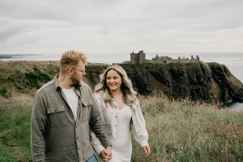 A couple walk hand in hand for engagement photos at Dunnottar Castle.