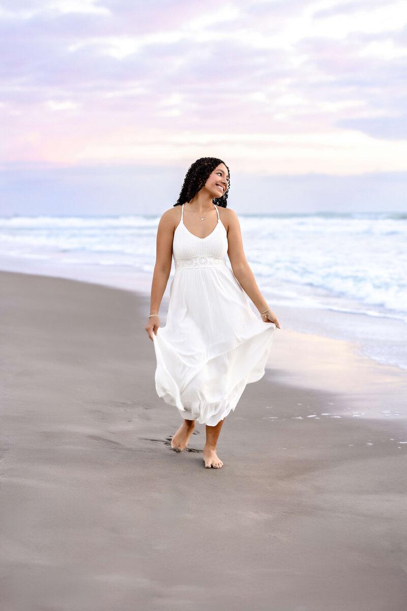 Teen girl in white dress walking barefoot along the shoreline at sunrise during an Orlando senior photo session at the beach.