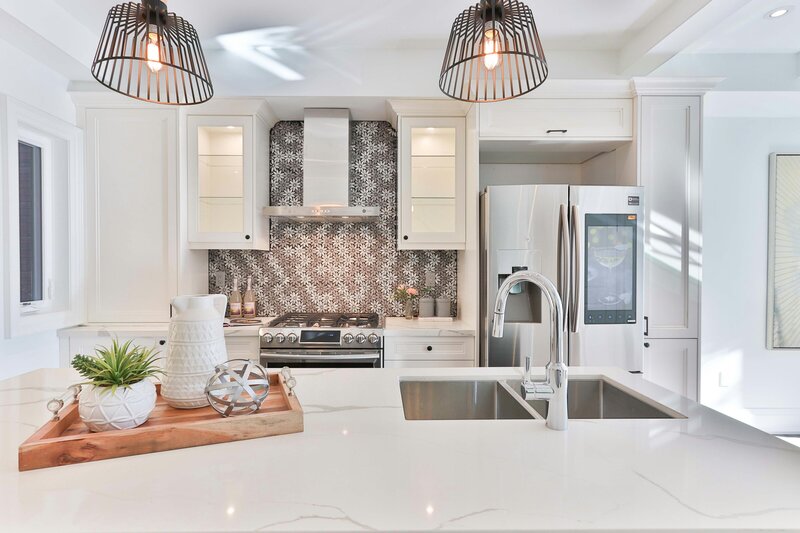 Close-up of a modern white kitchen with patterned tile backsplash, stainless appliances, glass cabinet, and quartz countertops.