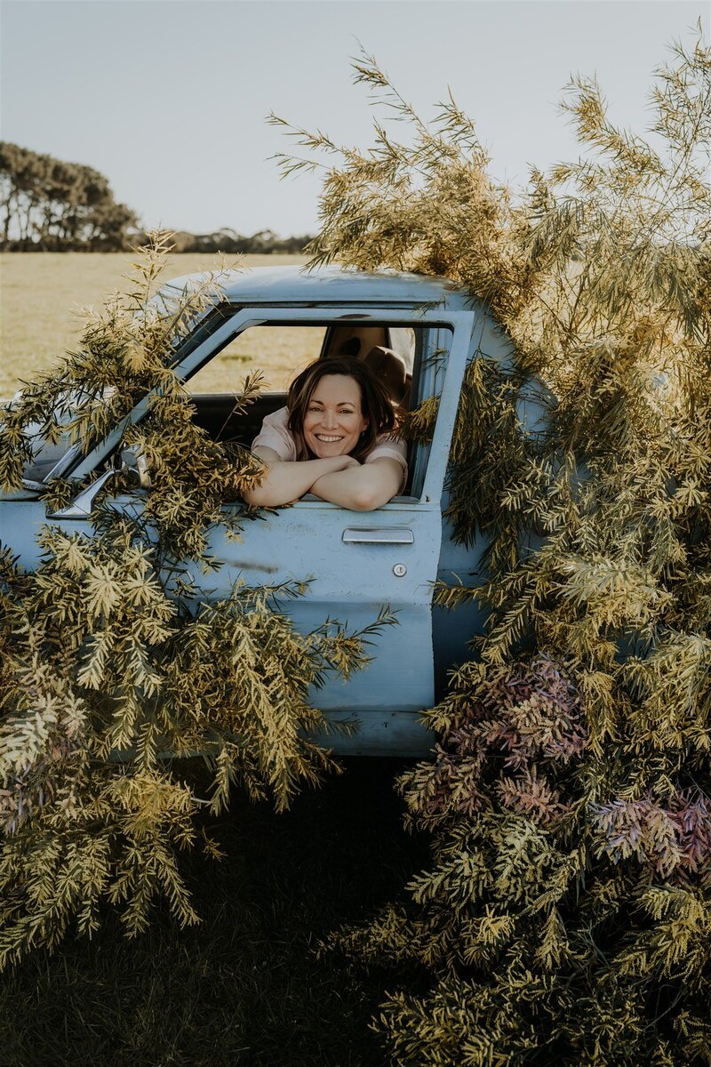 Picture of Corrie, the owner and florist behind Morrie & Maisie Event Flowers, sitting in a ute surrounded by wattle that she made for an editorial photoshoot