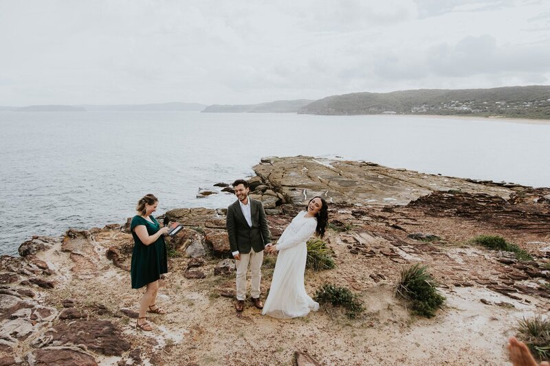 A couple are getting married in Bouddi National Park on their Elopement, there is a celebrant in the photos. They stand on a rocky cliff edge with a view behind them of the ocean, Killcare beach and a view to Sydney, Palm Beach and the Hawkesbury River in the distance. The image is shot from slightly above and the couple are laughing at something said by the celebrant.