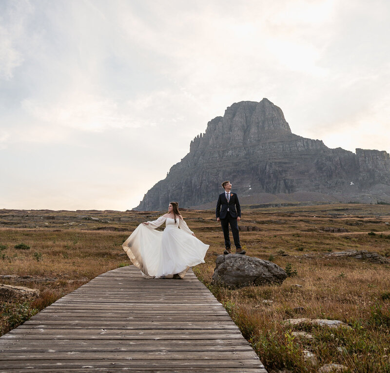 A bride twirls on a wooden boardwalk surrounded by golden fields and mountain cliffs during a Glacier National Park elopement, captured by Sydney Breann Photography