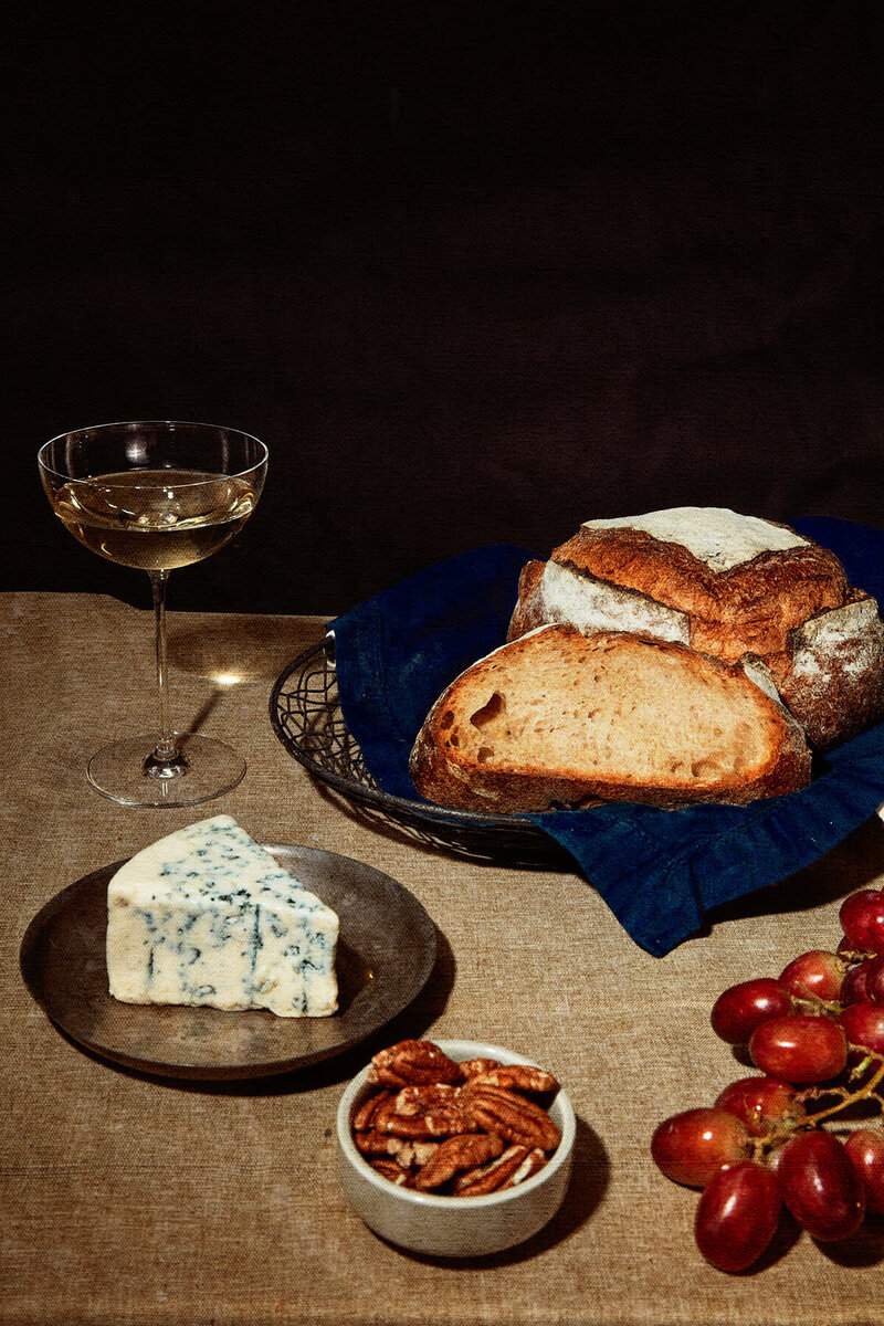 A classic still life featuring freshly baked bread, a wedge of blue cheese, pecans, and a glass of white wine. Captured by Mica McCook, this image exudes warmth and sophistication, perfect for showcasing the elegance of simple, artisanal foods.