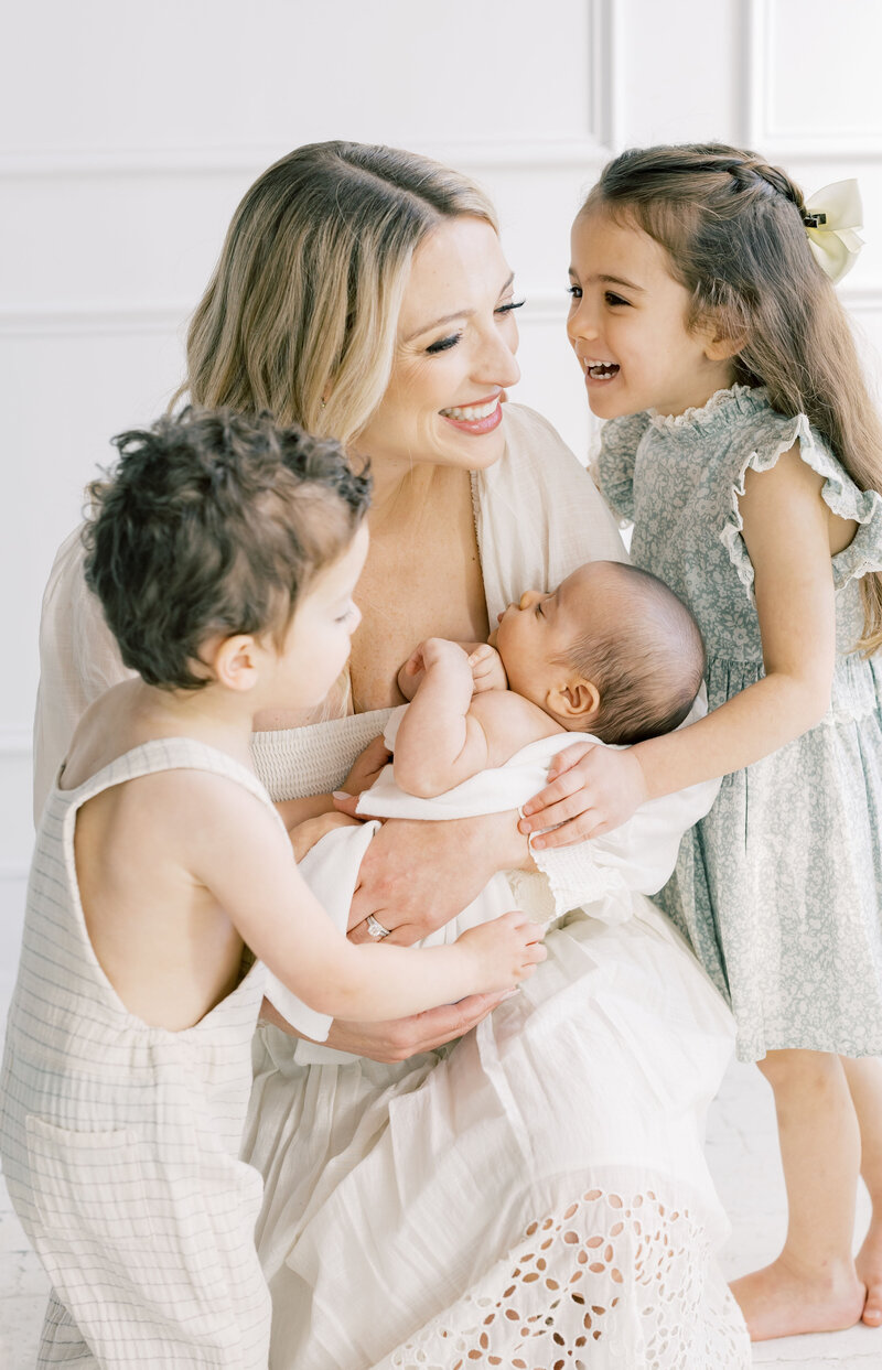 Mother embracing her daughters in a light-filled home, wearing coordinating neutral dresses during a lifestyle family session.