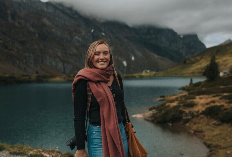 woman is smiling at the camera. She is wearing a pink chunky scarf, a black button up shirt. and jeans. She is smiling with a camera strap around her shoulder. there is a blue alpine lake behind her and mountains with snow and clouds.