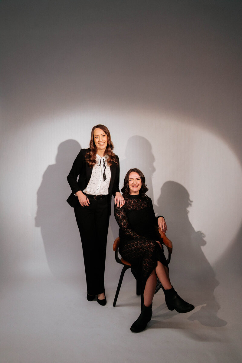 Two women pose in a studio spotlight background. One woman is standing, wearing a black suit an dwhite blouse, while the other is sitting on a chair wearing a black patterened dress and boots. Both are smiling. 