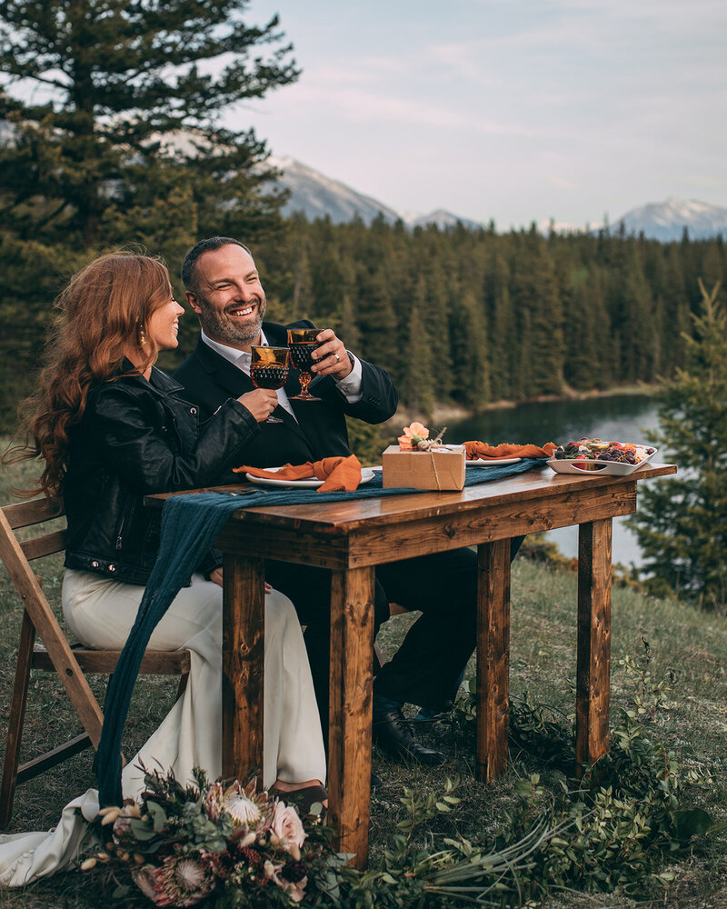 A couple sitting at a small table in the mountains toasting glasses during their elopement