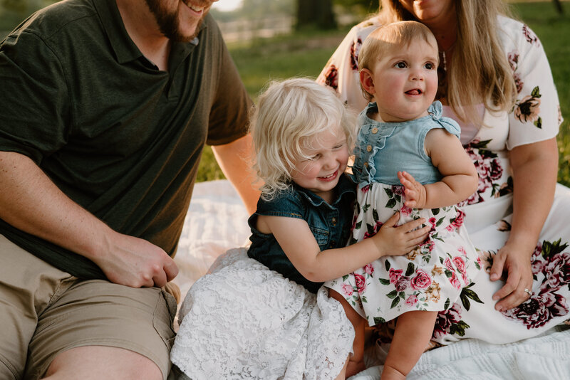 Family photo of mom, dad, and two little sisters on a blanket white older sister is going in to hug baby sister in Annapolis Maryland.