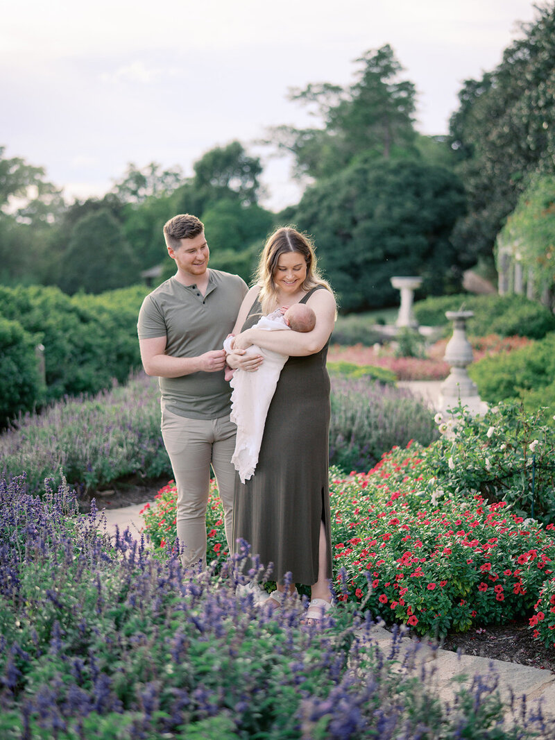 A mother standing in a garden holding a baby as the mother and father both look at the baby by Katie Stansfield Photography, a Richmond newborn photographer.