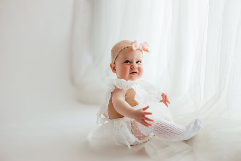 Seven month old baby girl sitting on a white floor with white sheer curtains dressed in a lace white dress is ready for her Denver milestone session