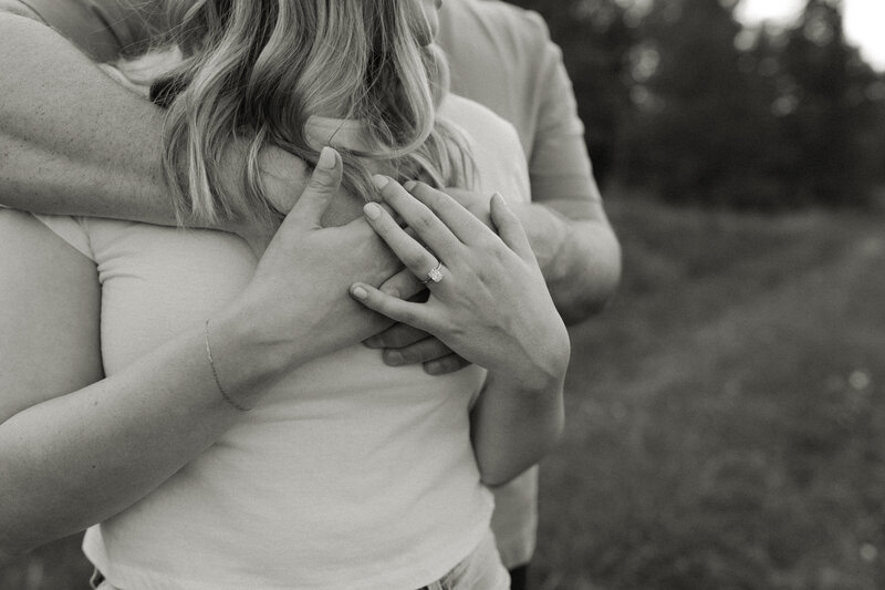 Man wraps his arms around his fiancé, who lovingly rests her hands over his while showing off her engagement ring.