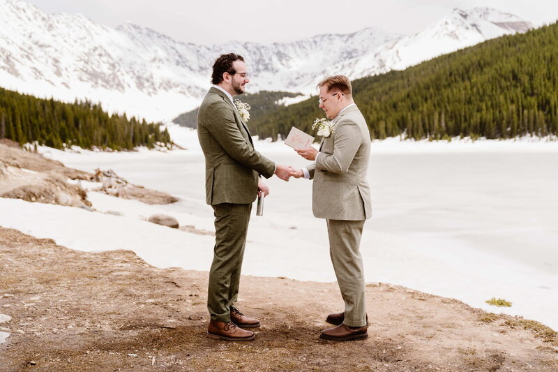 Two grooms exchange heartfelt vows by a frozen alpine lake in Leadville, Colorado, with snow-capped mountain peaks and evergreens in the background, photographed by Colorado elopement photographer Alpine Vows.