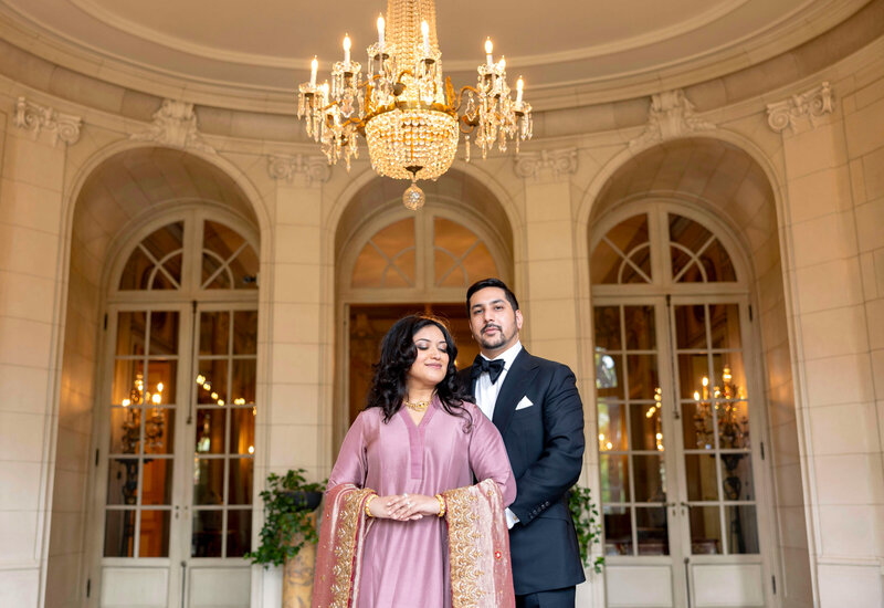 A couple stands together in a formal setting under a chandelier. The woman is wearing a traditional Pakistani outfit with intricate designs, while the man is in a tuxedo. They are in front of arched doorways with glass panels. The ambiance is elegant and classy.