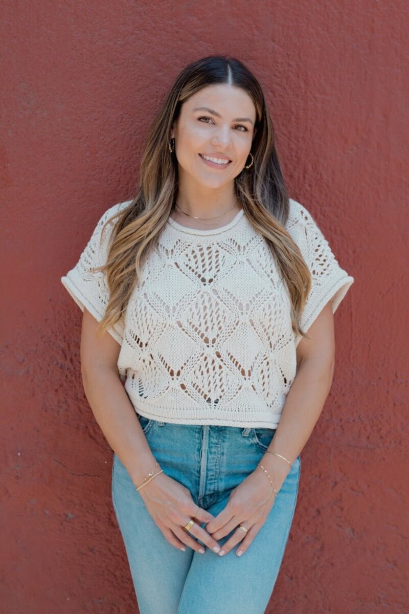 Natalie Bernacchi smiling on a bench by a cactus wall, wearing a straw hat and knit top with the text “Let’s create that shift together