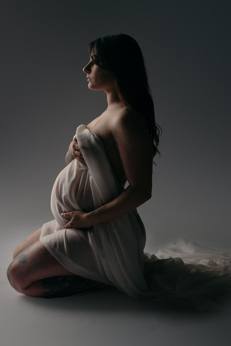 Pregnant woman nude wrapped in flowy chiffon fabric holding baby bump sitting on knees