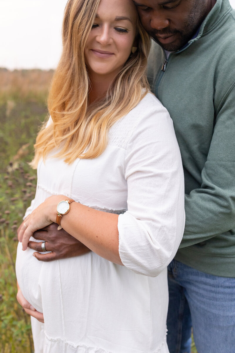 Expecting couple embraces in a natural outdoor setting, with the man wrapping his arms around the woman's baby bump. The woman smiles softly, wearing a white dress, while the man wears a green sweater and jeans.