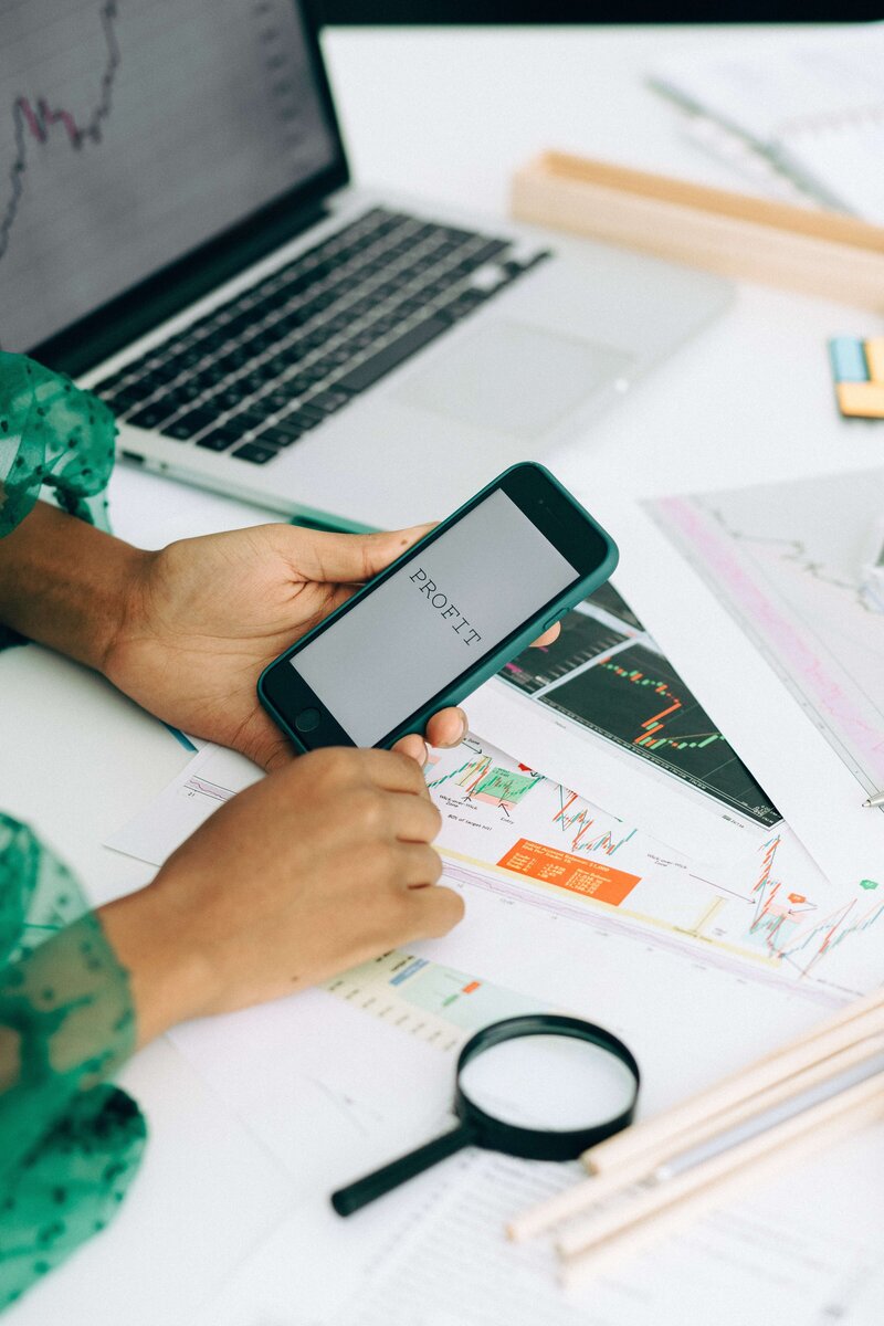 woman's hands holding a phone that reads "profit" above business charts