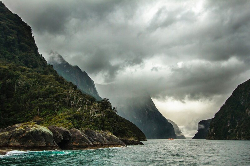 A moody image of Milford sound showing the rocky water edge and heavy grey clouds coming over the mountains.