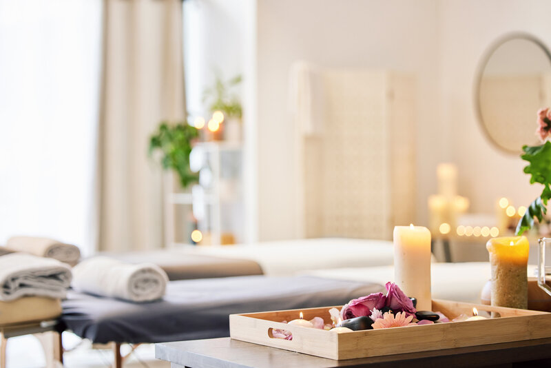 Close-up of spa essentials including bottles of massage oil, rolled towel, candles, and smooth stones on a wooden surface.