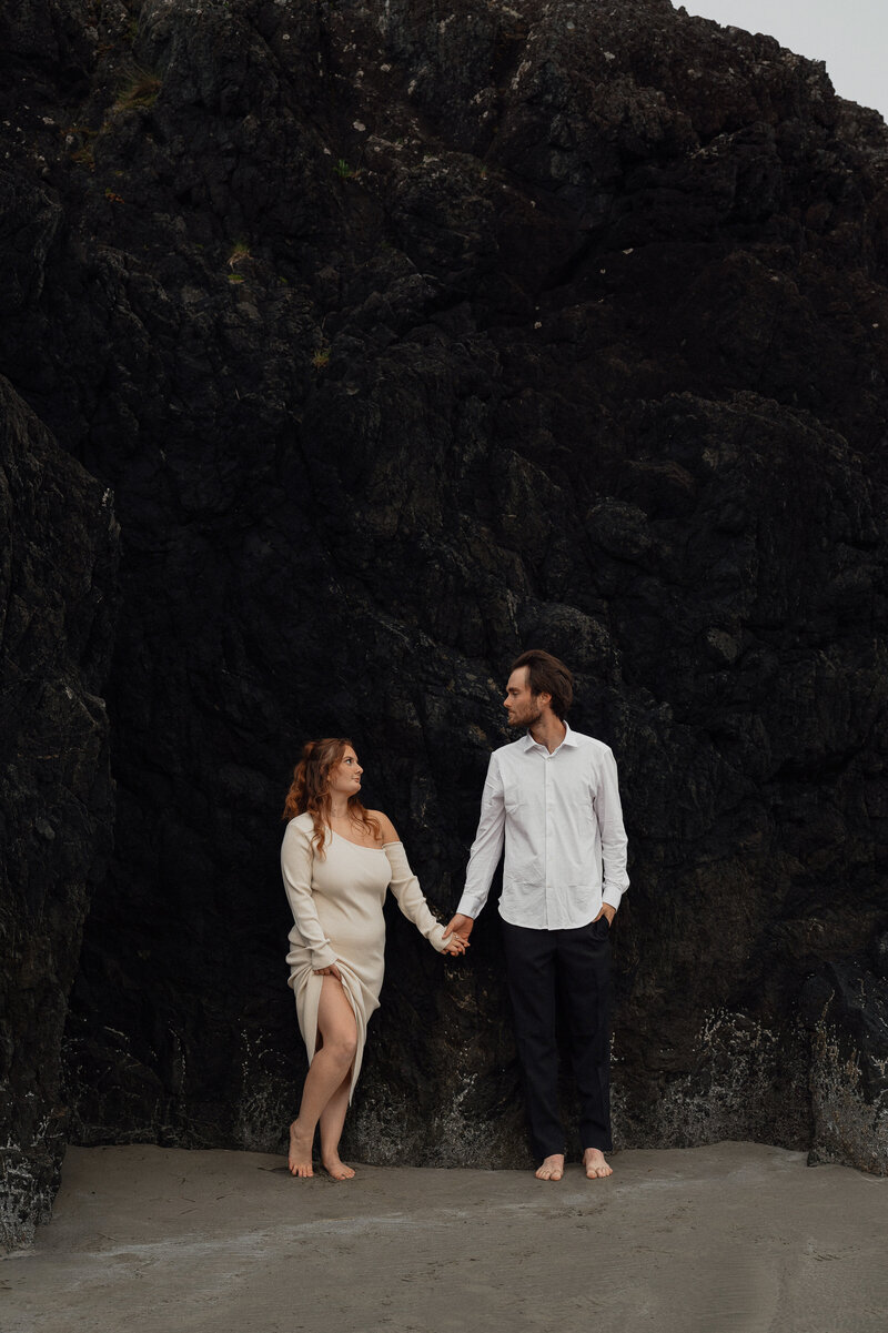 couple holding hands against the black rocks in Tofino at Long Beach  by Latitude 49 Photography.