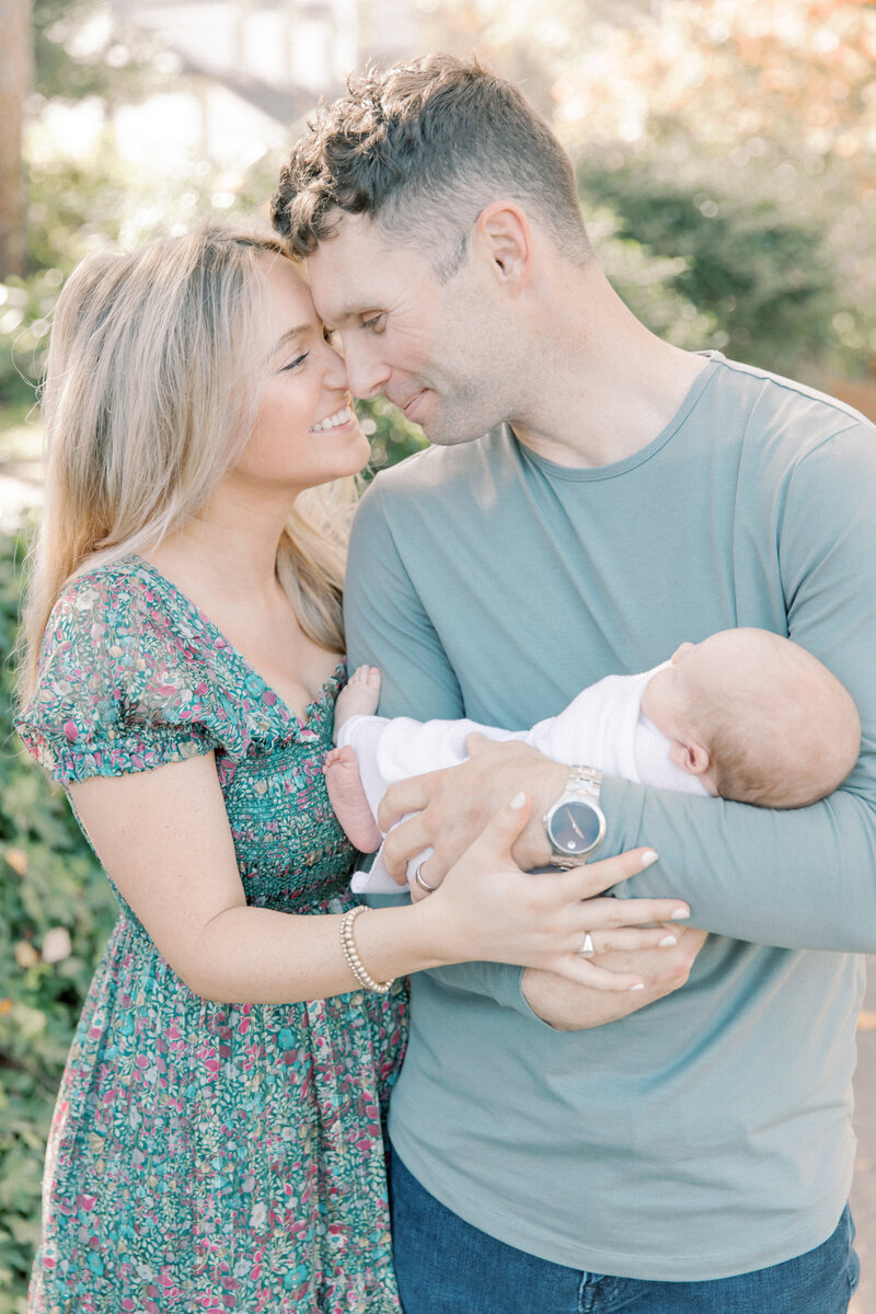 Parents holding their baby and smiling together outdoors in soft natural light.