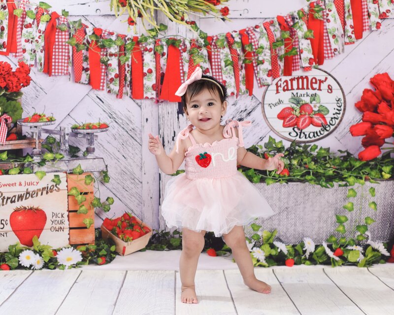 A baby girl standing in a pink tutu and strawberry-themed outfit, smiling in a Farmer’s Market setup with red and green decor.