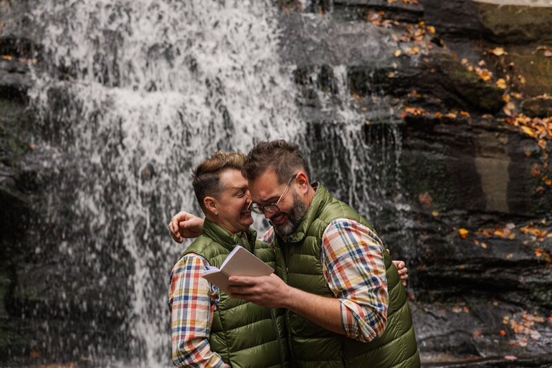 Two grooms are sharing an intimate moment and laugh while reading their vows to one another. They are standing in front of a waterfall flowing down rocks. 