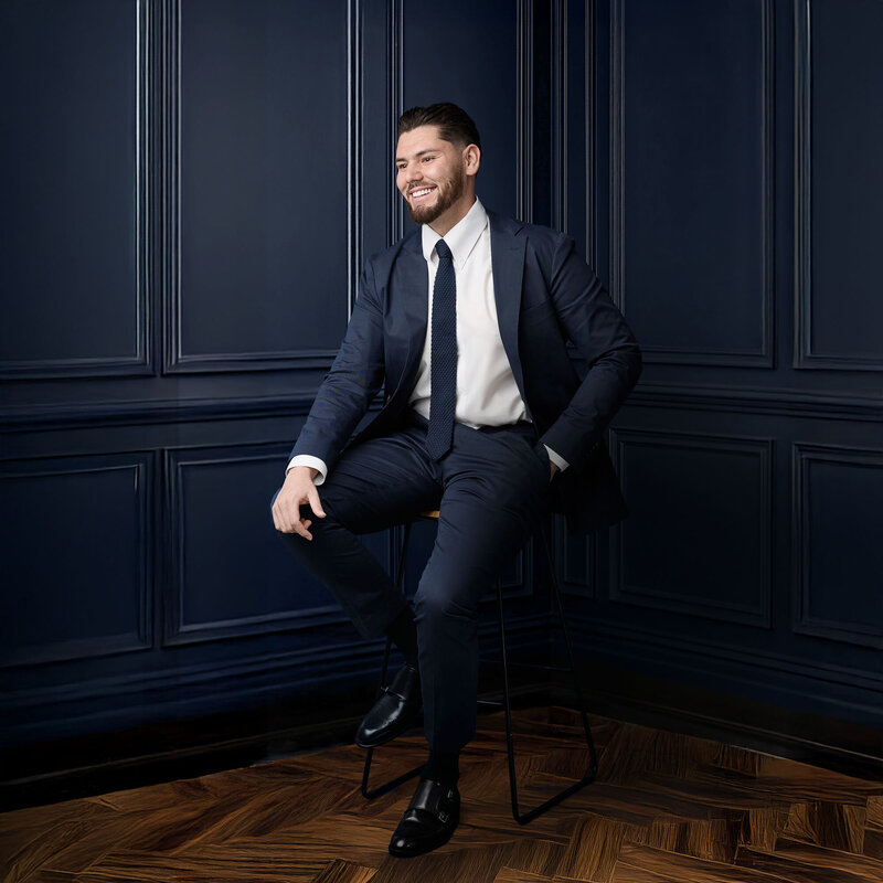 Stylish corporate man in a navy suit. He's on a stool against navy wainscoting and oak herringbone parquetry.