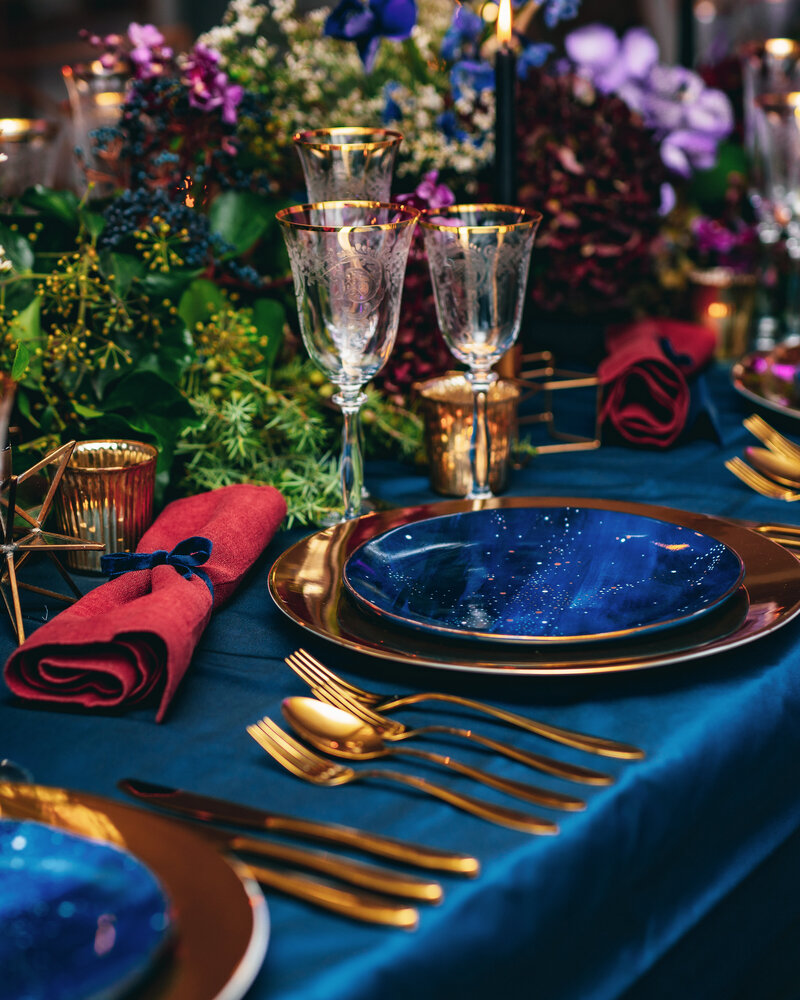 A celestial themed dinner table is set with a blue, gold and red accents amoung a luxurious display of flowers.