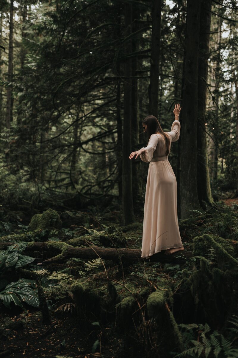 Woman stands in dark, lush forest wearing a cream-colored flowy dress with arms outstretched beside her as she balances on a horizontal tree.