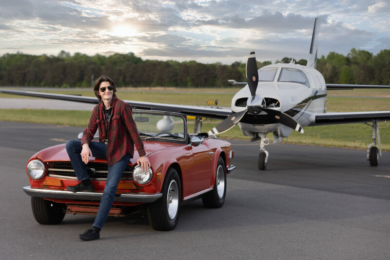 GRADUATING SENIOR SITTING ON THE HOOD OF HIS GRANDFATHERS OLD CONVERTIBLE CAR WITH AN AIRPLANE BEHINE THE CAR.  BEAUTIFUL IMAGE OF HIS FAVORITE THINGS!