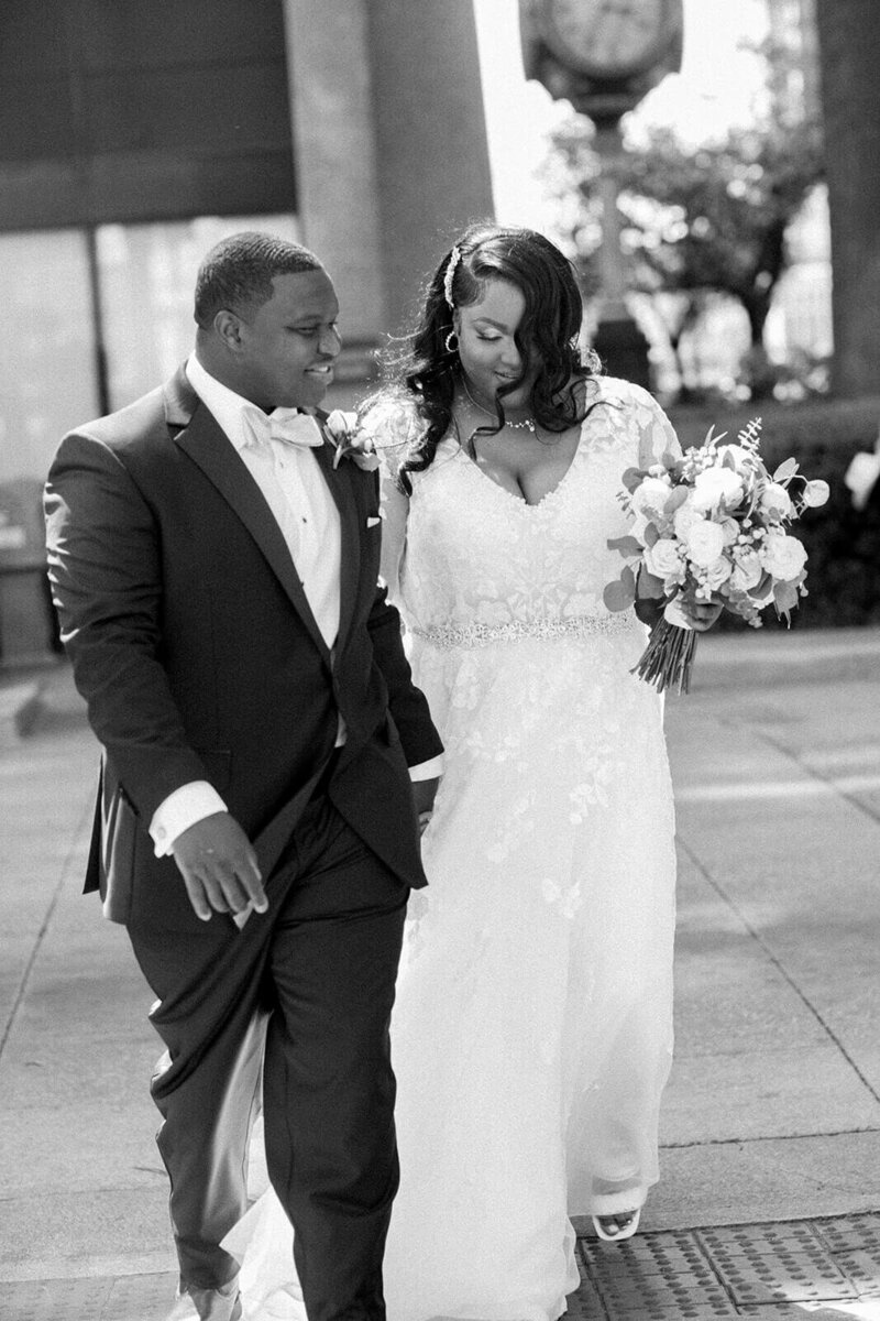 A Black bride and groom walk hand in hand outside their wedding venue, 1208 Washington in Columbia, SC. The groom wears a classic black tuxedo with a white bow tie, smiling as he looks at his bride. The bride, in a lace wedding gown with a beaded belt, carries a bouquet of white and blush flowers while gazing downward. A vintage clock and cityscape frame the background in this timeless black-and-white wedding portrait.
