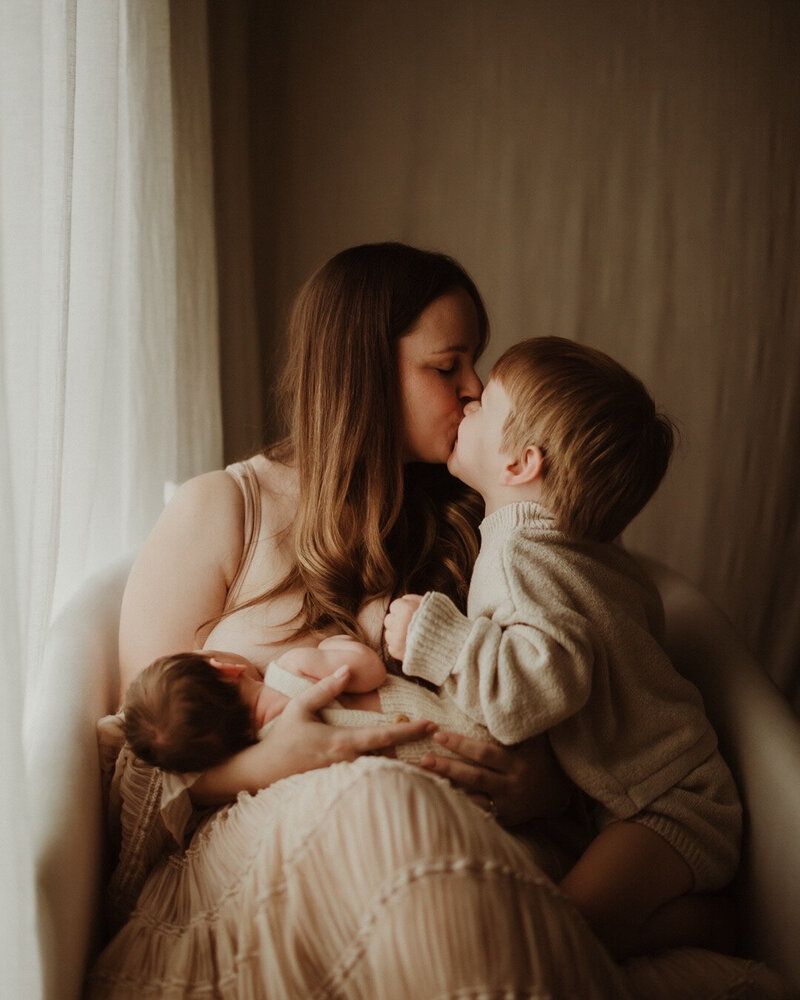 Mother breastfeeding baby in warm natural light while giving son a kiss, taken by Sydney motherhood photographer Sophie Lea.