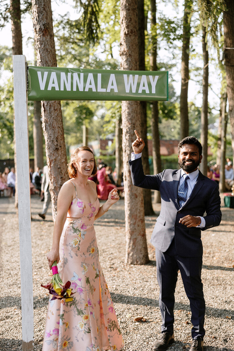 A woman in a floral dress holds a bouquet, while a man in a suit gestures next to a sign reading “Vannala Way” amidst trees.