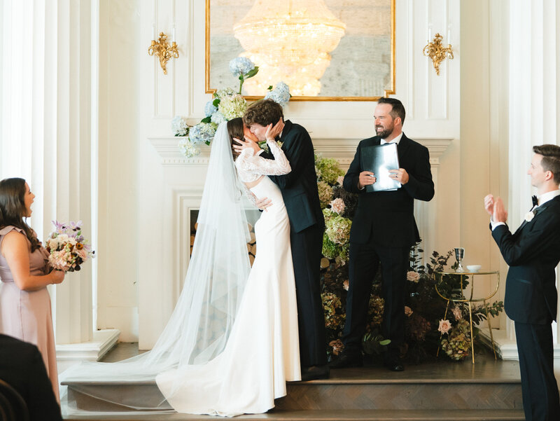 Bride and groom kissing on stage at their ceremony while bridal party cheers