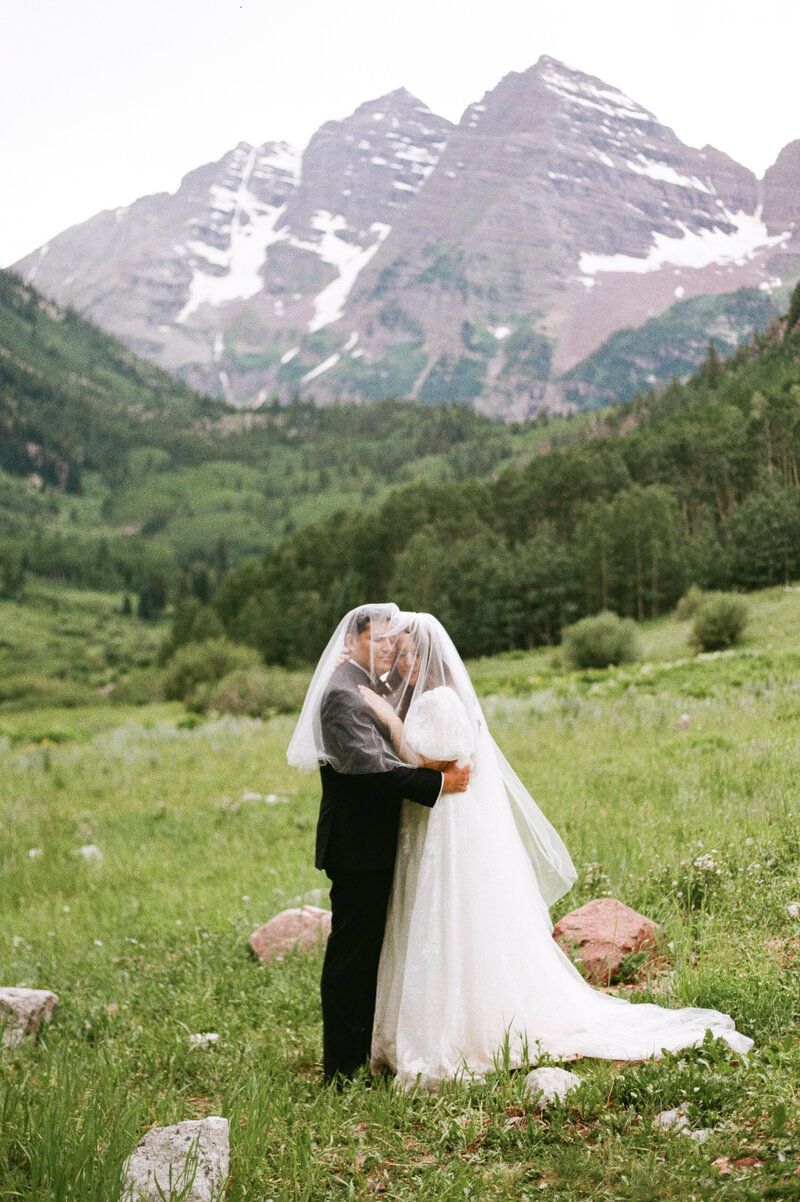Couple embraces under a shared veil in front of the Maroon Bells Amphitheater wedding, surrounded by wildflowers and alpine scenery captured by Fort Collins, Colorado portrait photographer Avenir Photo Co.