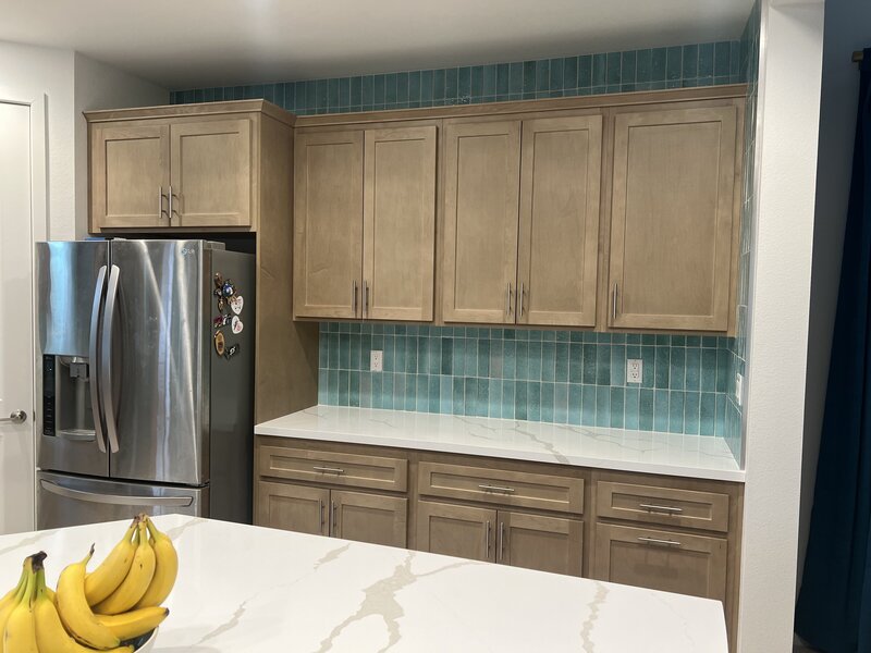 A remodeled kitchen with light wood cabinets, a teal vertical tile backsplash that goes to the ceiling, and faux white marble counter tops.