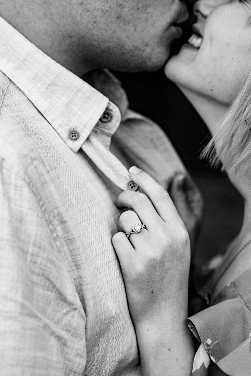 Black and white detail photo of the bride's diamond halo engagement ring.  The bride holds the groom's shirt as she shows off her ring while leaning in for a kiss. She is smiling up at the groom.