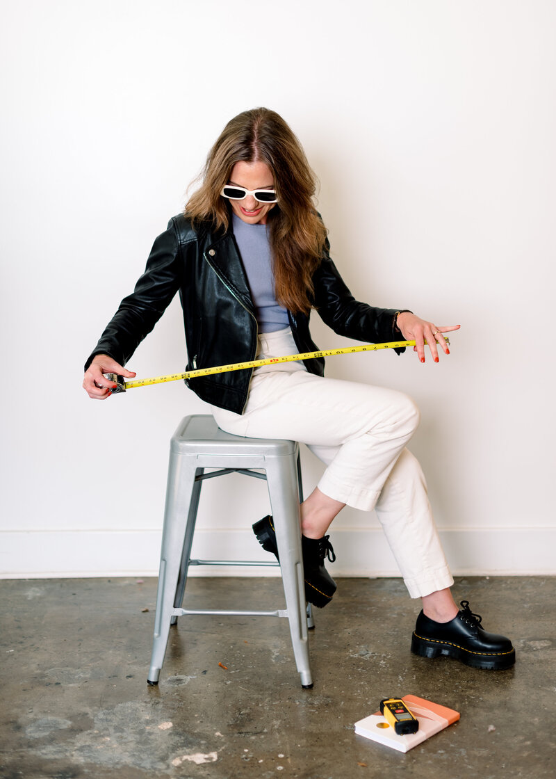 durham interior designer sitting on metal stool with tape measure, doc martens, leather jacket, and white sunglasses inspired by kelly wearstler