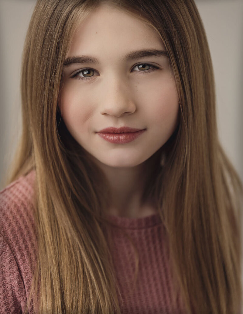 A young person with straight, long brown hair wears a textured pink top. They look directly at the camera with a calm, neutral expression.