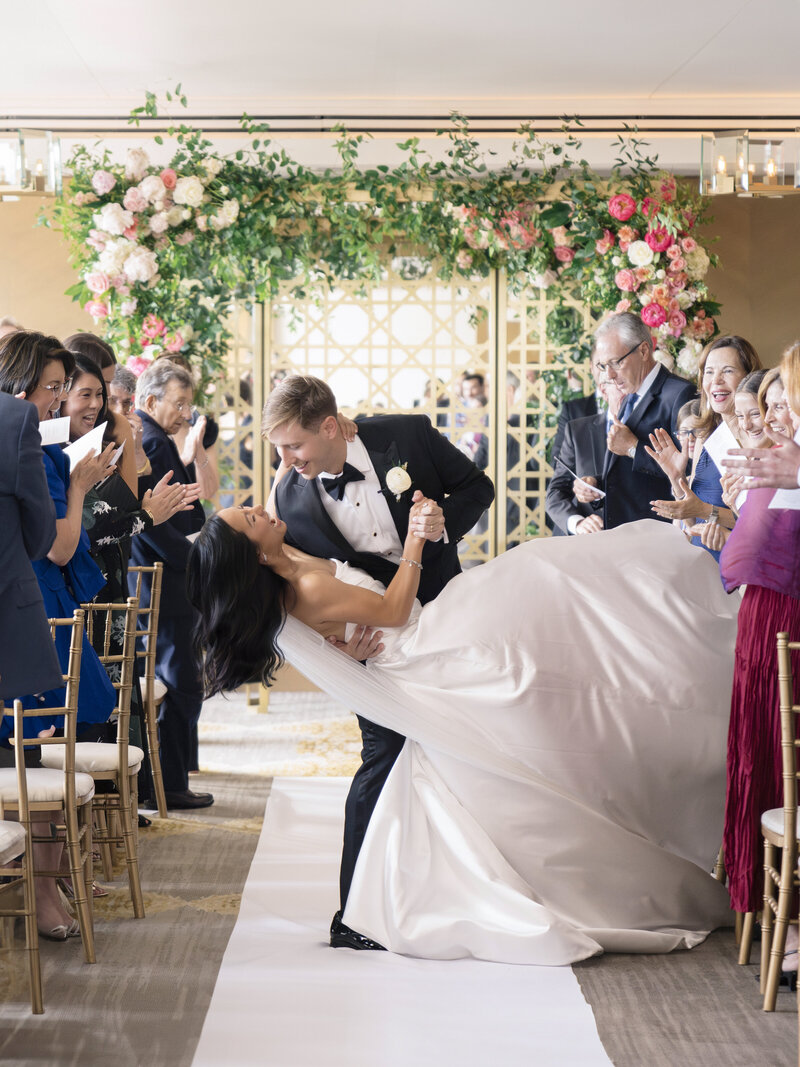 A newlywed couple shares a kiss in an elegantly decorated wedding aisle. The groom, in a black tuxedo, dips the bride, who wears a flowing white gown. Guests clap and smile, surrounded by floral arrangements and gold accents.
