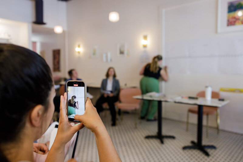 At a networking event in Seattle, a person in the foreground is taking a photo with a smartphone of two women in a bright, modern room. One woman is sitting with a notepad, while the other writes on a large sheet of paper on the wall.