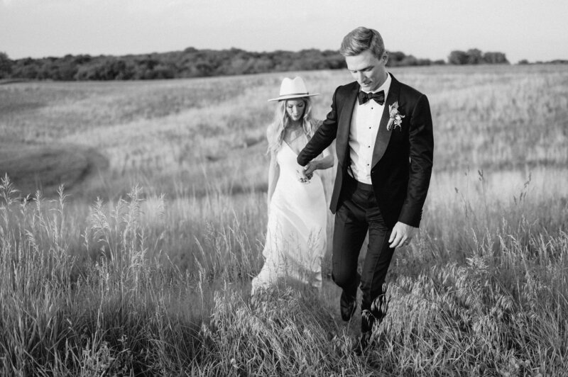 Bride wearing a hat and groom wearing a black tux walk up a hill holding hands. Photo by Anna Brace, a wedding photographer in Omaha Nebraska