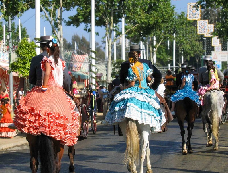 Horse riders in traditional Feria outfits - men in black suits and hats with women in coral and turquoise ruffled flamenco dresses seated behind them - at the April Fair in Seville, Spain ©Stephanie Dosch | theViatrix Luxury Travel