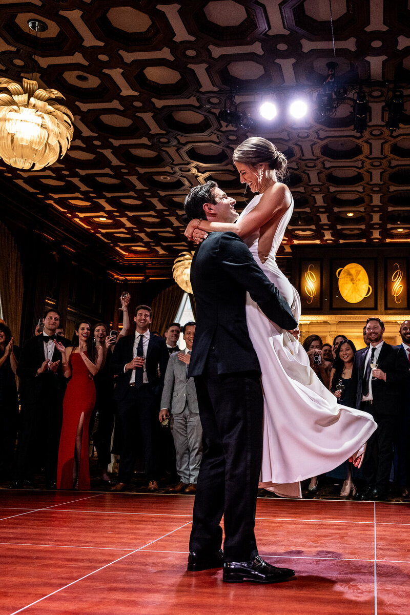 San Francisco Bay Area wedding photographer captures intimate first dance as groom lifts bride at Julia Morgan Ballroom with intricate coffered ceiling and celebrating guests