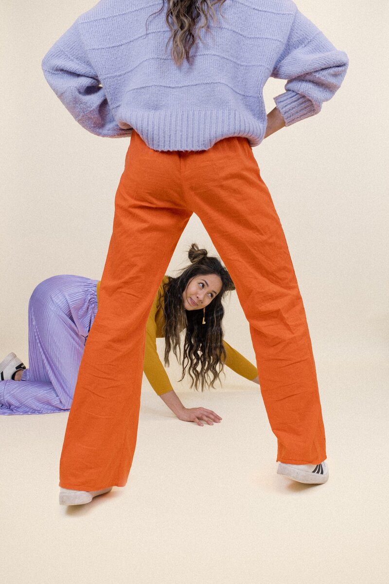 Woman with half-up pigtail buns crawls on the floor in front of a photoshopped duplicate version of herself wearing a purple top and orange pants with her back to the camera.