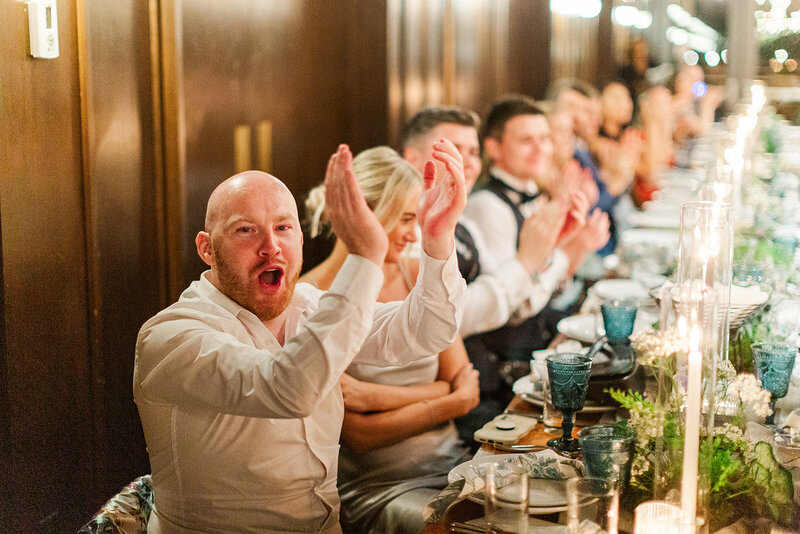 Wedding guests seated for dinner a long farm table in the ballroom at District Winery in DC clapping and cheering